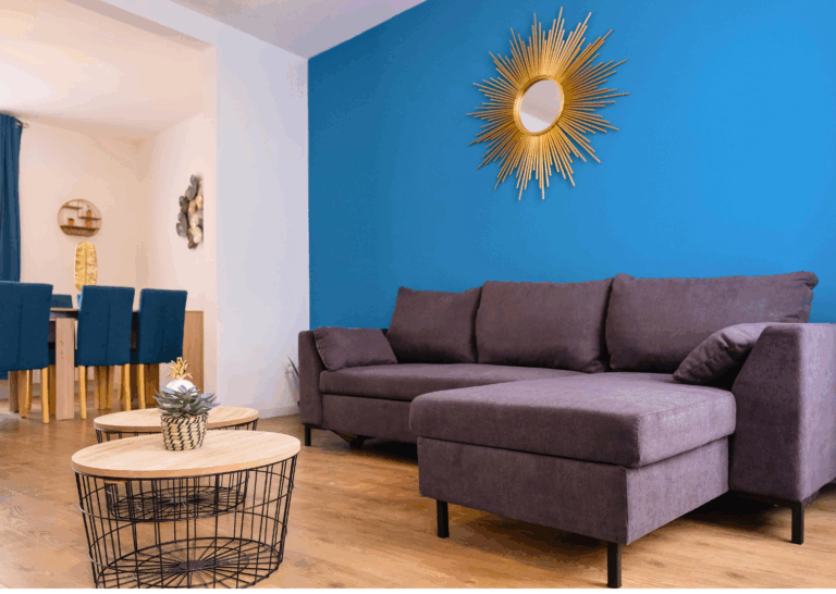Salon moderne avec canapé d’angle gris, tables basses en bois et mur bleu décoré d’un miroir soleil doré – hébergement Apart’Moment à Saint-Omer.